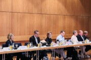 An einer Reihe von Konferenztischen mit Getränken, Mikrofonen und Namensschilder darauf sitzen Menschen in Business Kleidung.