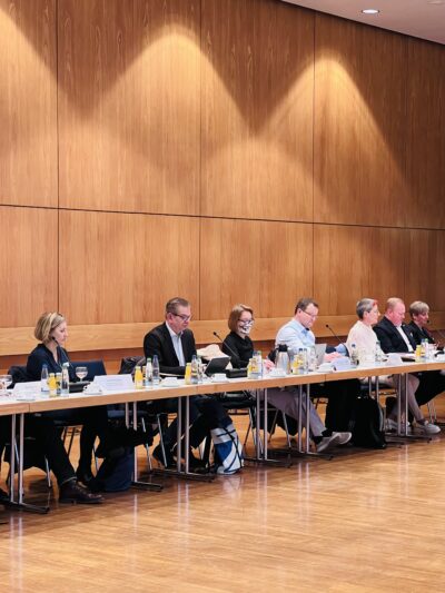An einer Reihe von Konferenztischen mit Getränken, Mikrofonen und Namensschilder darauf sitzen Menschen in Business Kleidung.