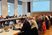 Das Bild zeigt einen langen Sitzungstisch mit Männern und Frauen, die aufmerksam zuhören oder ihre Unterlagen studieren.